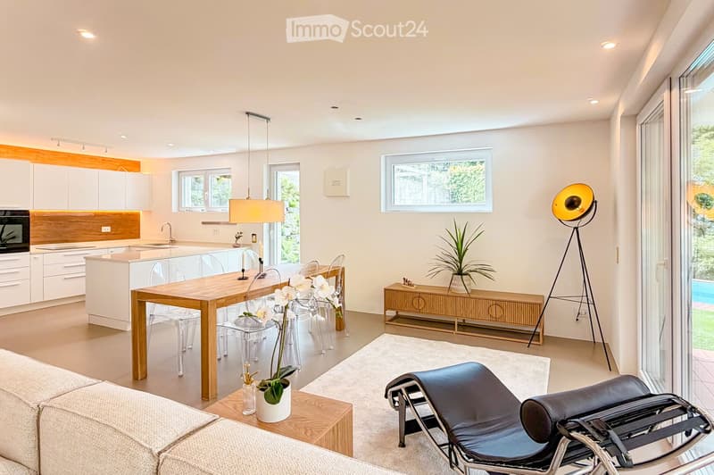 Furnished Room. Modern open-plan living and dining area with a white kitchen, wooden dining table, transparent chairs, a black leather lounge chair, and a floor lamp.