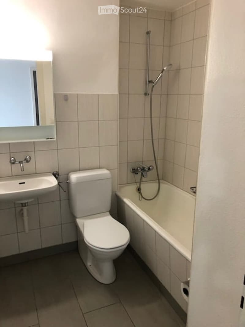 Modern bathroom with white tiles, a toilet, a sink, a bathtub with a shower hose, and a mirror above the sink.