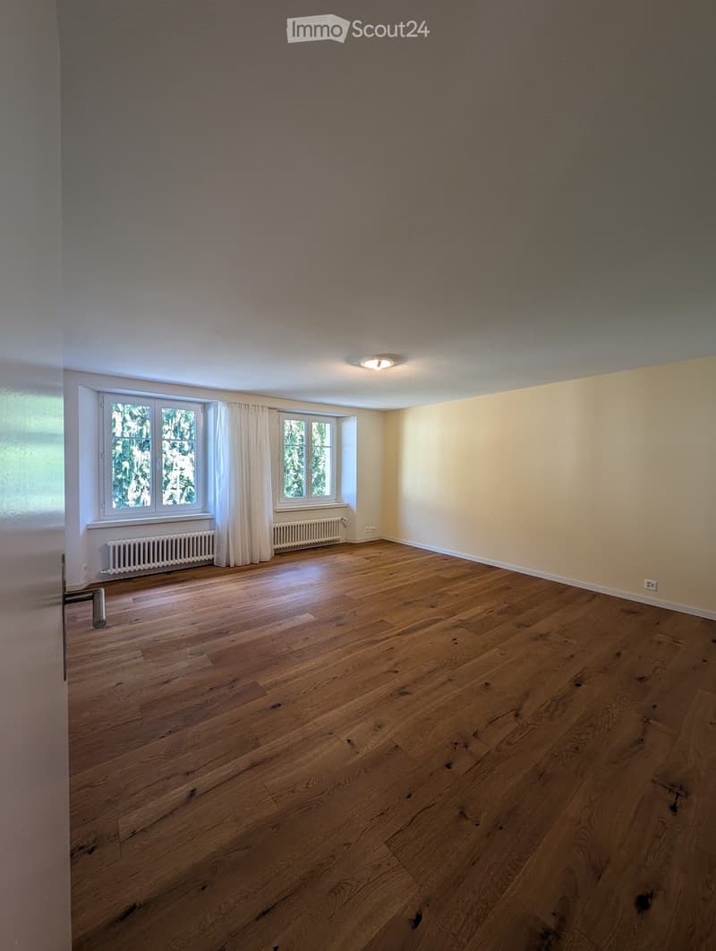 Empty room with wooden flooring, large windows with white curtains, and a radiator beneath the windows.