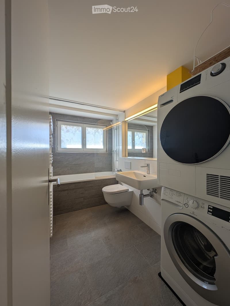 Modern bathroom with a toilet, sink, bathtub, and washing machine. Features include a large window, sleek fixtures, and a clean, contemporary design.