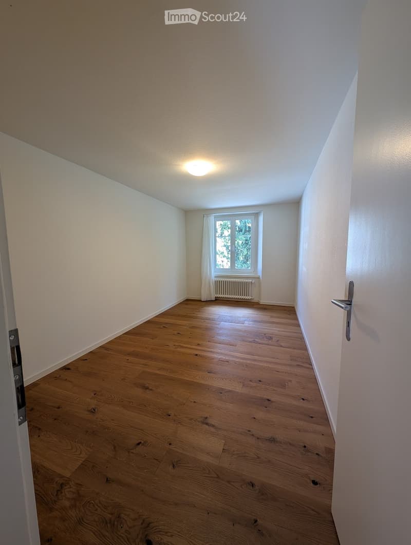 Empty room with wooden flooring, white walls, a window with white curtains, and a radiator below the window. The room is well-lit with a ceiling light fixture.