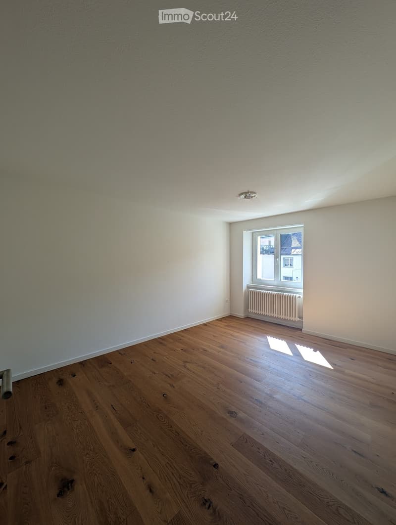 Empty room with wooden flooring, white walls, and a window with a radiator below it. The room is well-lit with natural light coming through the window.