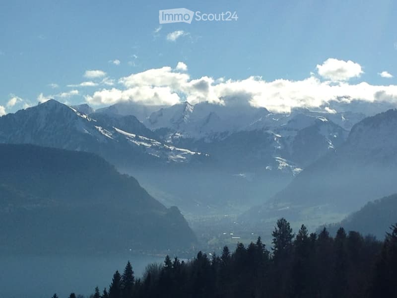 A panoramic view of snow-capped mountains and a forested valley under a partly cloudy sky.