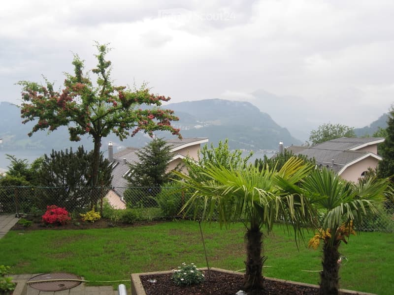 Well-maintained garden with green grass, flowering trees, shrubs, and palm plants, overlooking a scenic lake and mountain view.