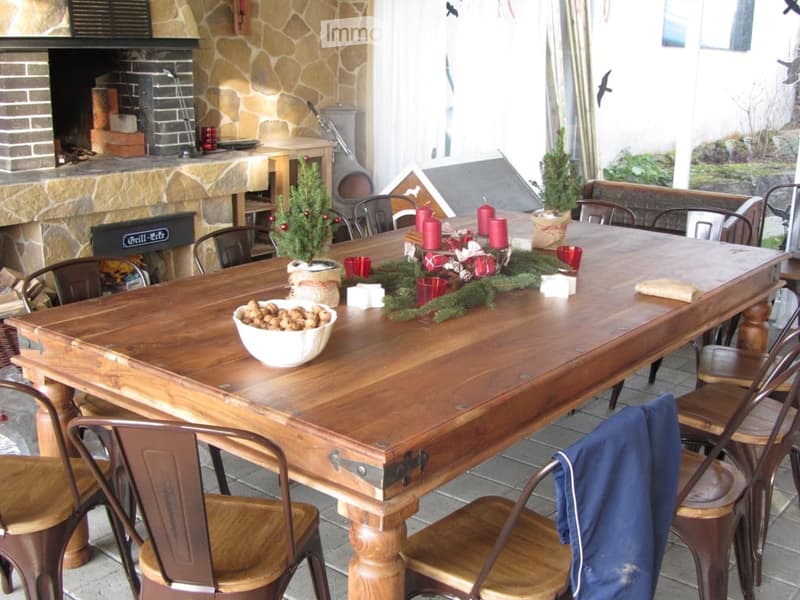 An outdoor dining area with a large wooden table, surrounded by wooden chairs, a fireplace, and festive decorations including candles and a small Christmas tree.