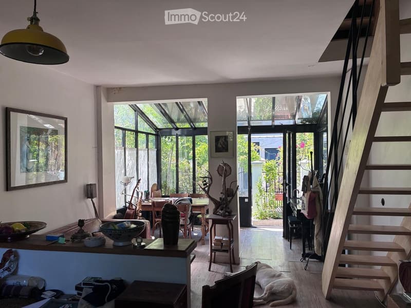 Bright living room with large windows, wooden floor, sofa, table, chairs, decorative items, and a staircase leading upstairs.