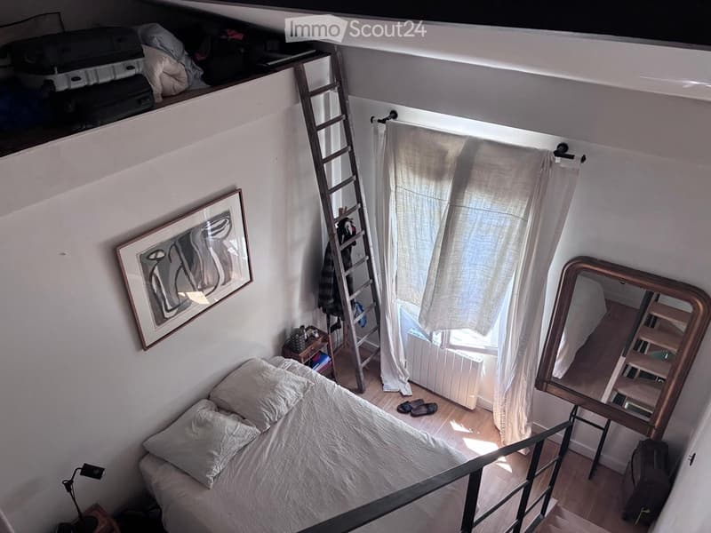 Cozy attic bedroom with a single bed, wooden floor, large window with white curtains, ladder to loft area, and a small table with a lamp.