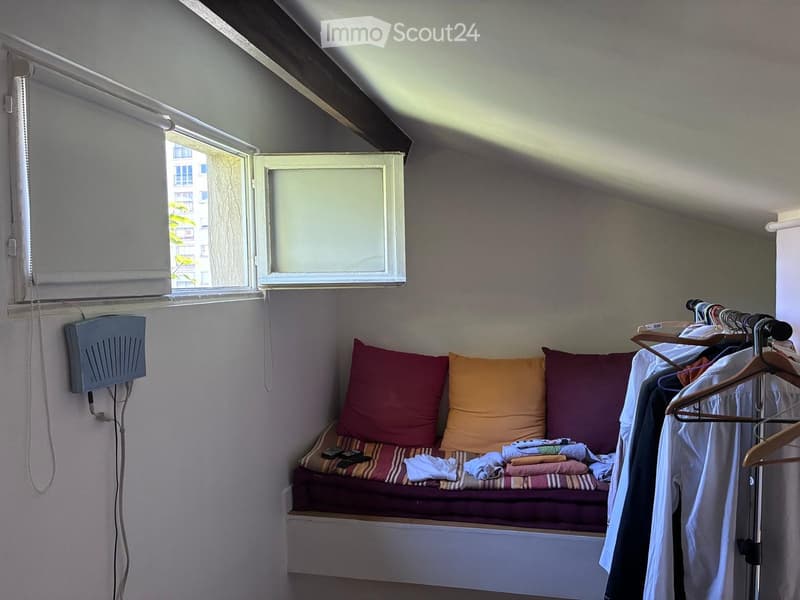 Small, cozy attic room with a built-in sofa, colorful cushions, a remote control, and a clothing rack with hanging clothes.