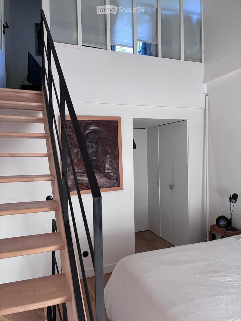Modern bedroom with a white bed, wooden nightstand, digital clock, and a staircase leading to another level.
