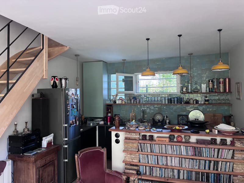 Modern kitchen with a central island, open shelving, and a staircase leading to an upper level. The space features a refrigerator, sink, and various decorative items on shelves and surfaces.