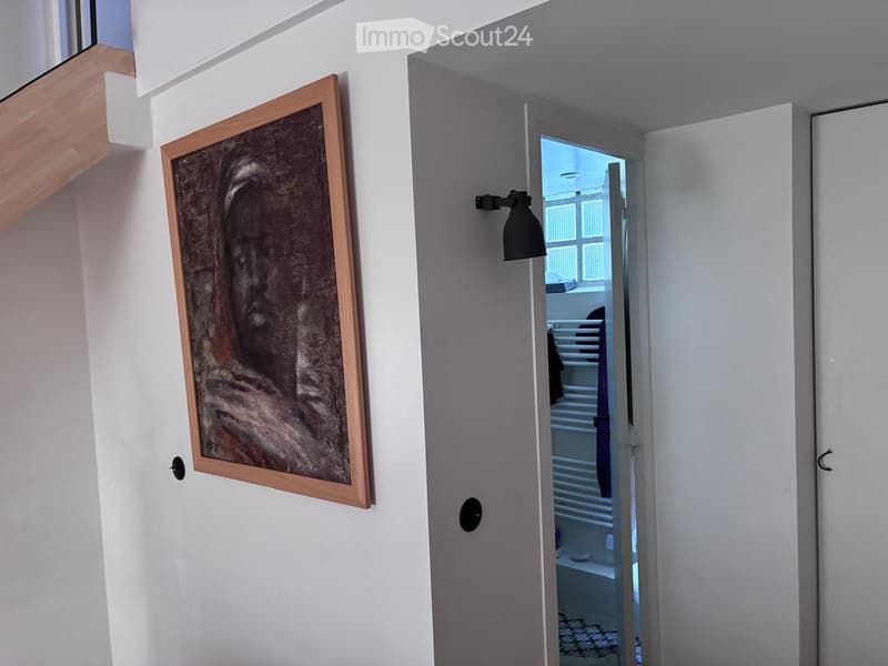 A narrow hallway with white walls, a framed portrait on the left, and an open door leading to another room with visible furniture.