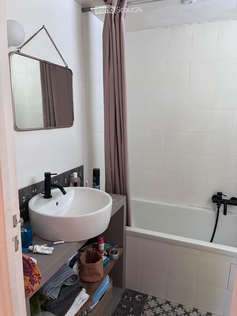 Modern bathroom with a white sink, black faucet, round mirror, bathtub, and various toiletries on the counter.