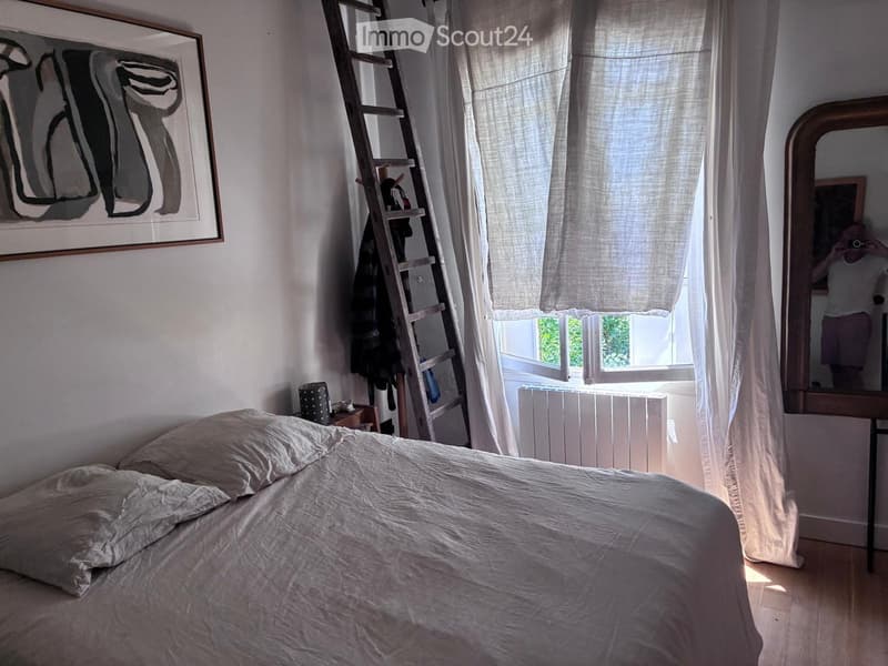 Cozy bedroom with a large bed, nightstand, mirror, and artwork on the wall. Natural light filters through sheer curtains.