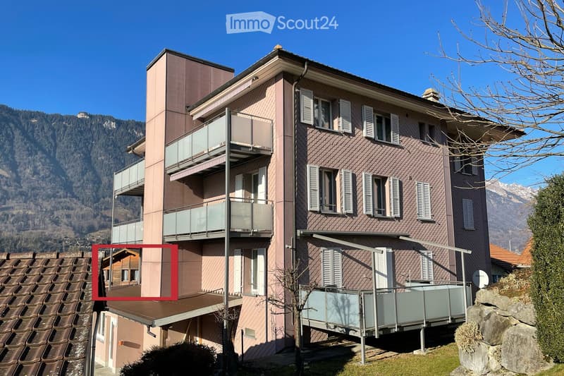 A modern two-story building with a brown tiled roof, multiple windows with shutters, and balconies with glass railings. The building is situated in a scenic area with mountains in the background.