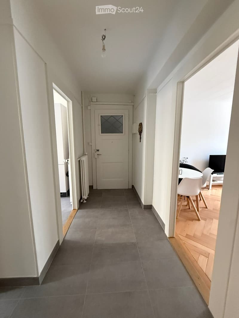 A narrow hallway with grey tiled flooring, white walls, and a central door leading to another room. The hallway has a radiator on the left wall and a small decorative item on the right wall. The hallway leads to a room with wooden flooring, white chairs,