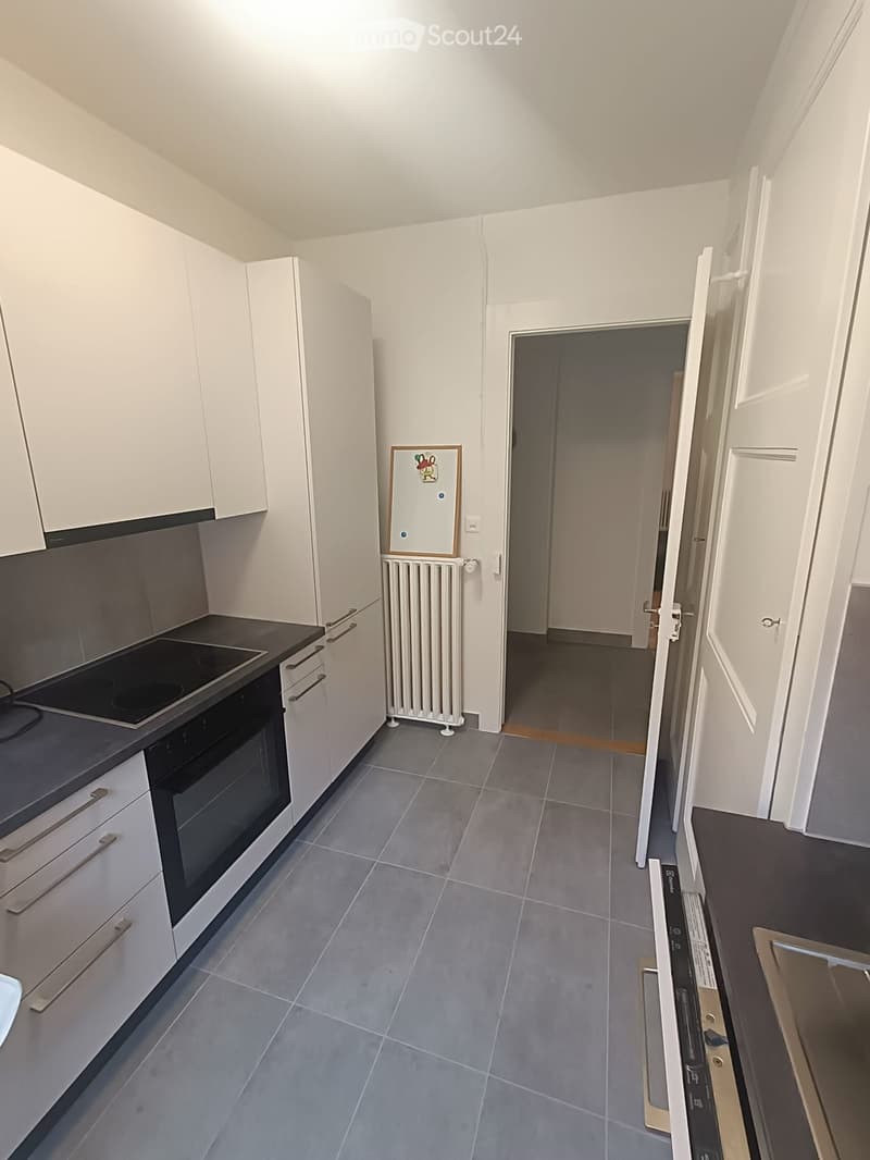 Modern kitchen with white cabinets, grey tiles, and an oven. A radiator is visible near the wall, and a doorway leads to another room.