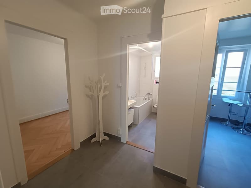 A modern hallway with wooden flooring, white walls, and a coat rack. Visible rooms include a bathroom with a bathtub and a room with blue flooring and bar stools.