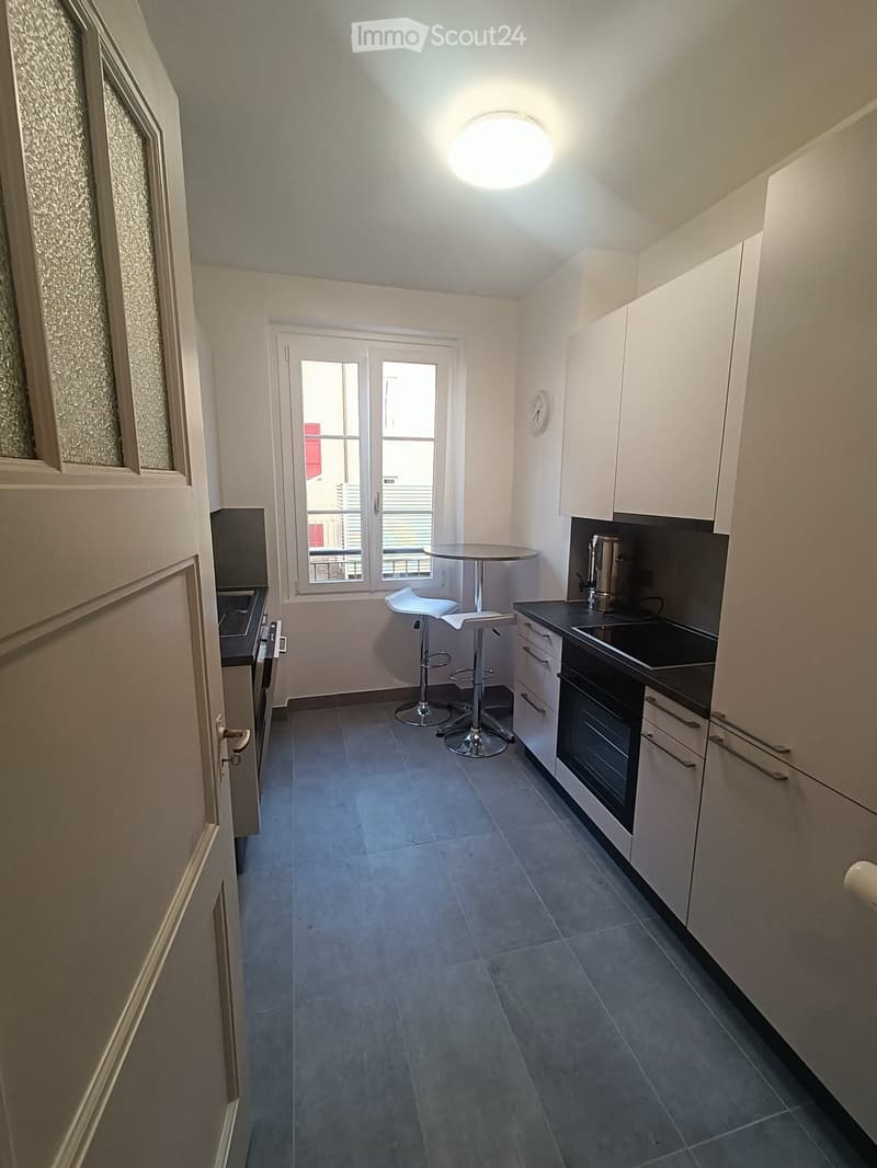 Modern kitchen with white cabinets, grey tiled floor, round table with two stools, oven, and refrigerator.