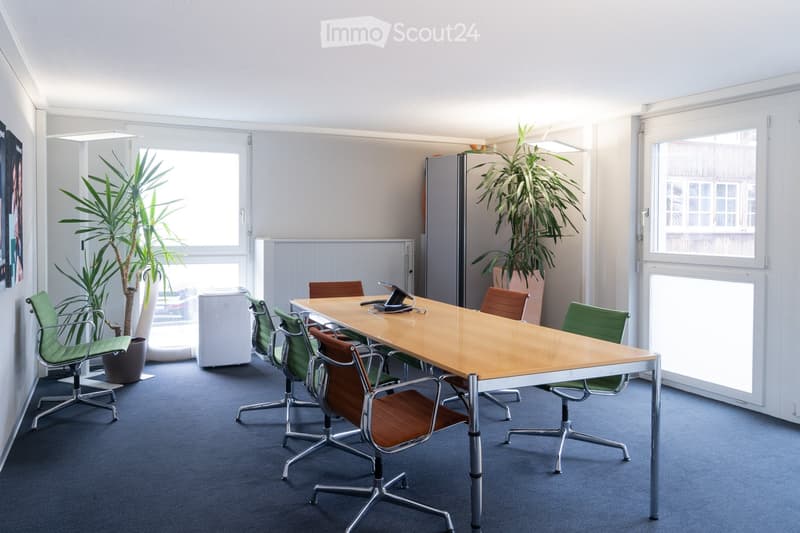 A modern office space with a large wooden conference table surrounded by green and brown chairs, potted plants, and a refrigerator in the background.