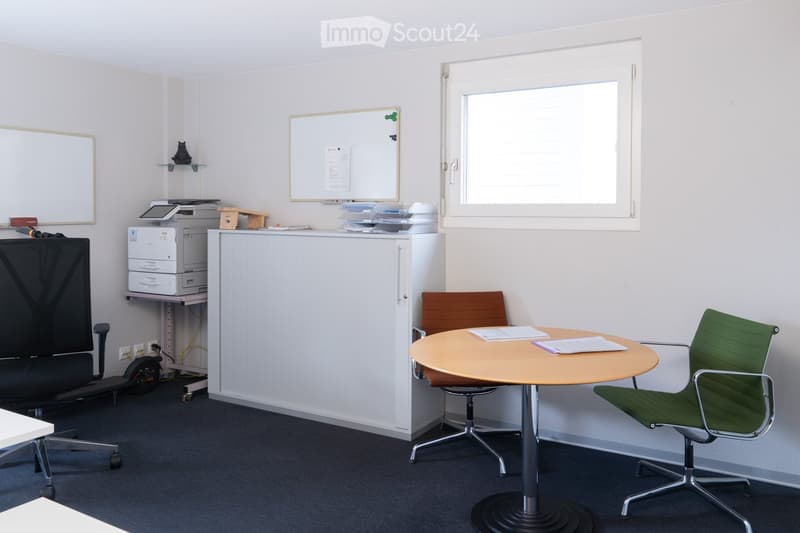 Office space with a round table, two chairs, a cabinet, a printer, and a screen. There are also shelves with various items and a whiteboard on the wall.
