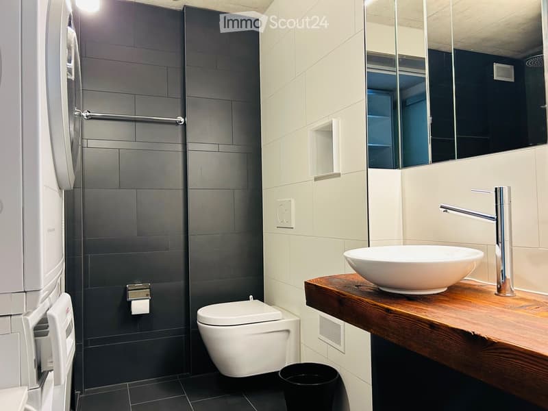 Modern bathroom with white toilet, wall-mounted white sink, black tiled floor, and wooden countertop. There is a washing machine on the left side and a trash bin on the right.