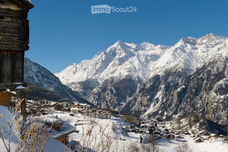 Mountainous landscape with a snow-covered village in the foreground, traditional wooden buildings, mountains in the background, clear blue sky.