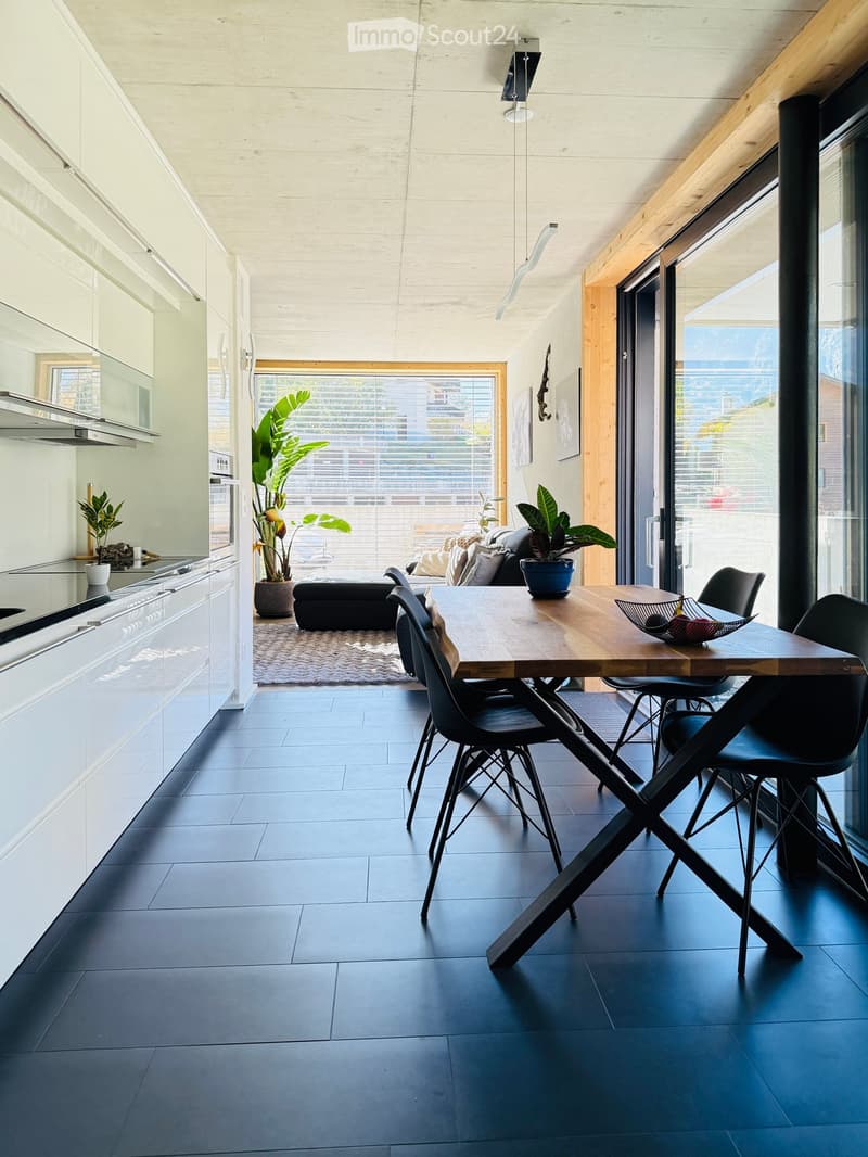 Modern kitchen with black tiles, white cabinets, open dining area with table and chairs, glass door to living room with couch and potted plants, large window with blinds.