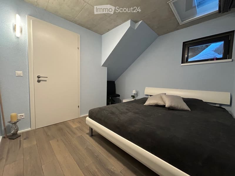 Bedroom with a large bed, wooden floor, white walls, night light, window, and nightstand with plant and candle.