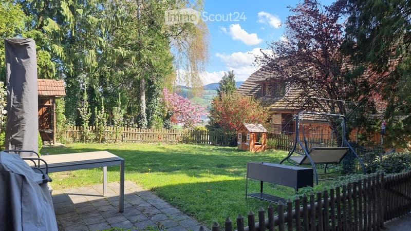 Garden with a swing, table, and covered furniture, surrounded by trees and a wooden fence.