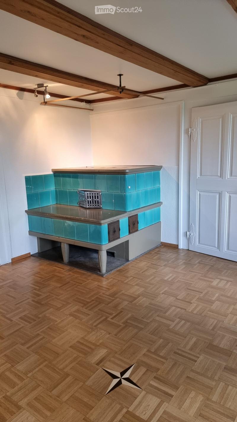 Empty room with wooden herringbone flooring, white walls, exposed wooden beams on the ceiling, and a fireplace with teal tiles and a metal grate. A white door is visible on the right side.