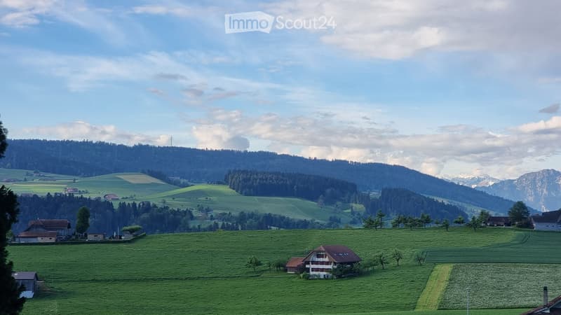 A picturesque rural landscape with green fields, scattered houses, and distant mountains under a partly cloudy sky.
