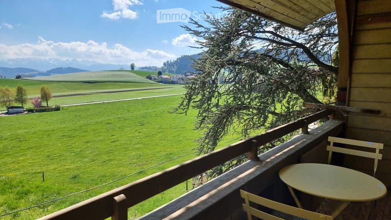 Balcony with wooden railing, overlooking a green field and distant mountains, with a small table and chairs.