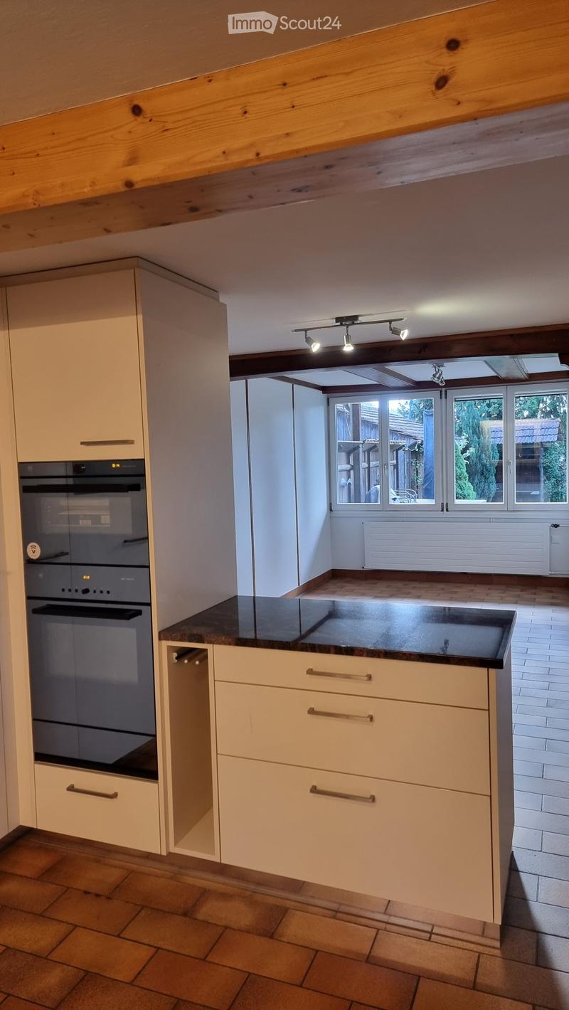 Modern kitchen with light-colored cabinets, a dark countertop, and an integrated oven. The space features exposed wooden beams on the ceiling and large windows offering a view of the outdoor area.