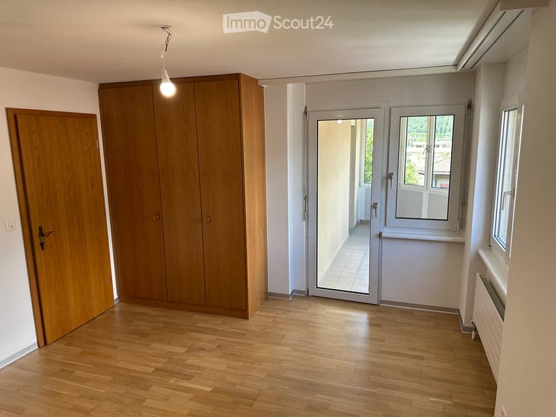 Empty hallway with wooden flooring, white walls, and a large wooden wardrobe. There are two doors, one leading to another room, and a window with a view of the outside.