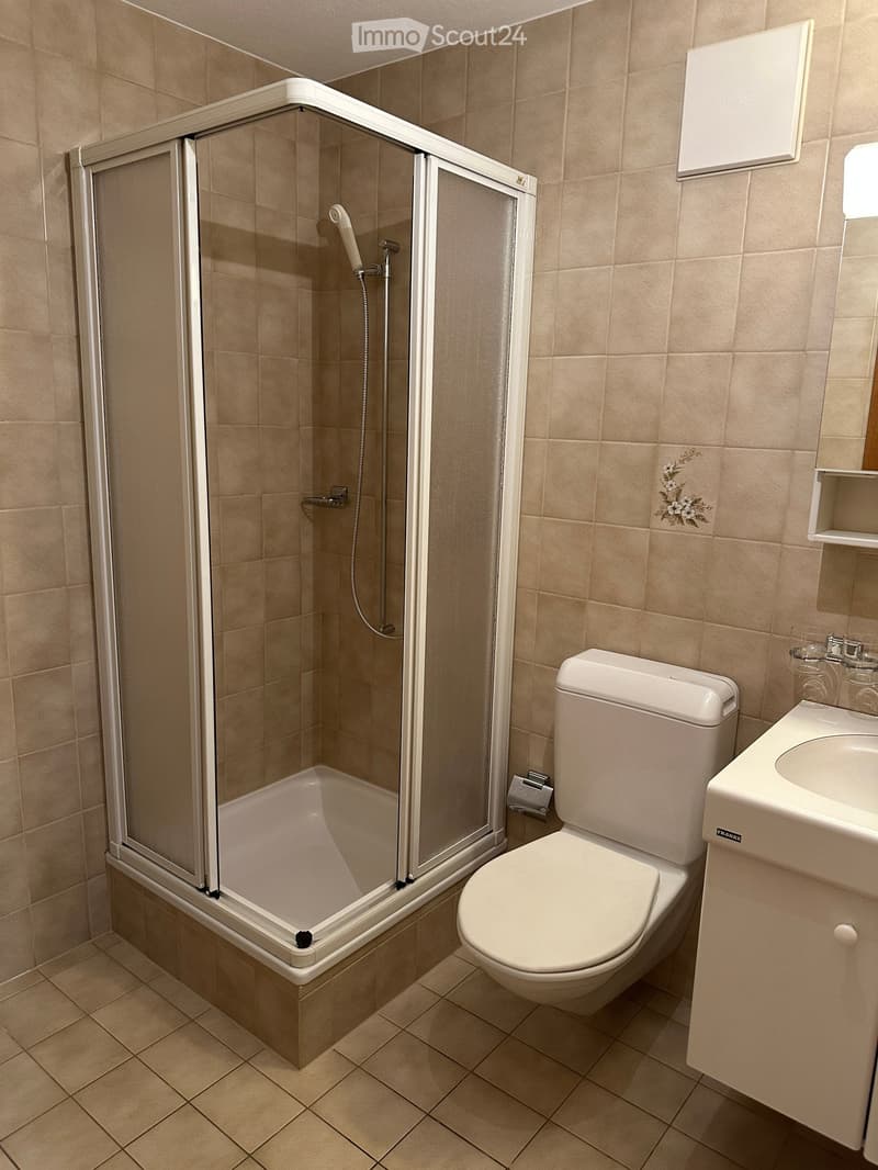 Modern bathroom with beige tiles, a corner bathtub with a glass shower enclosure, a toilet, and a sink with a mirror above it.