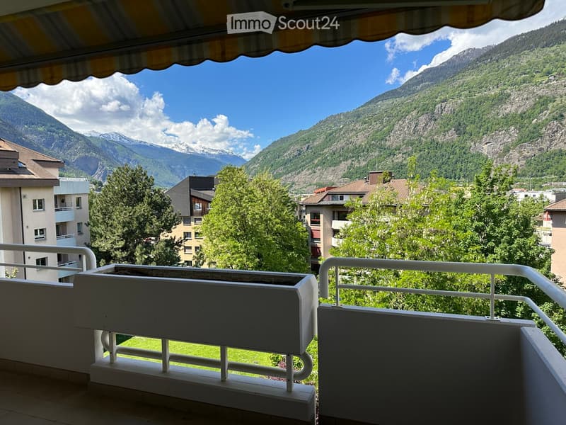 Balcony with white railings, overlooking a scenic mountain view and surrounding buildings.