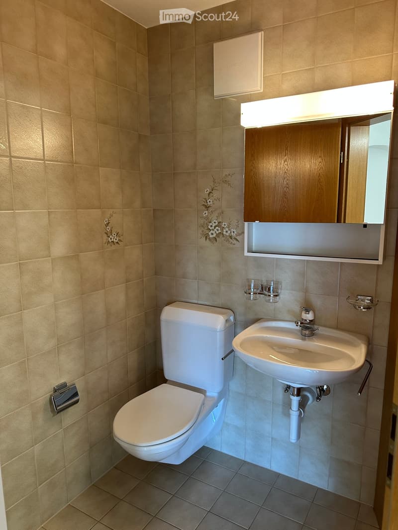 Modern bathroom with beige tiled walls and floor, featuring a toilet, sink, mirror, and wooden cabinet with a built-in light fixture above it.