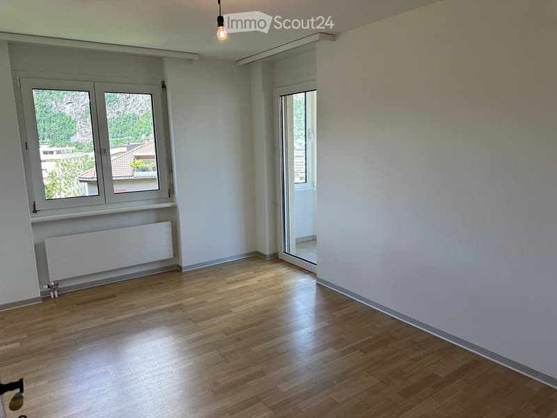 Empty room with wooden flooring, white walls, large windows, radiator, and a hanging light fixture.