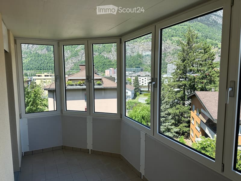 Balcony with large windows offering a scenic view of a town and surrounding mountains, tiled floor, white walls.