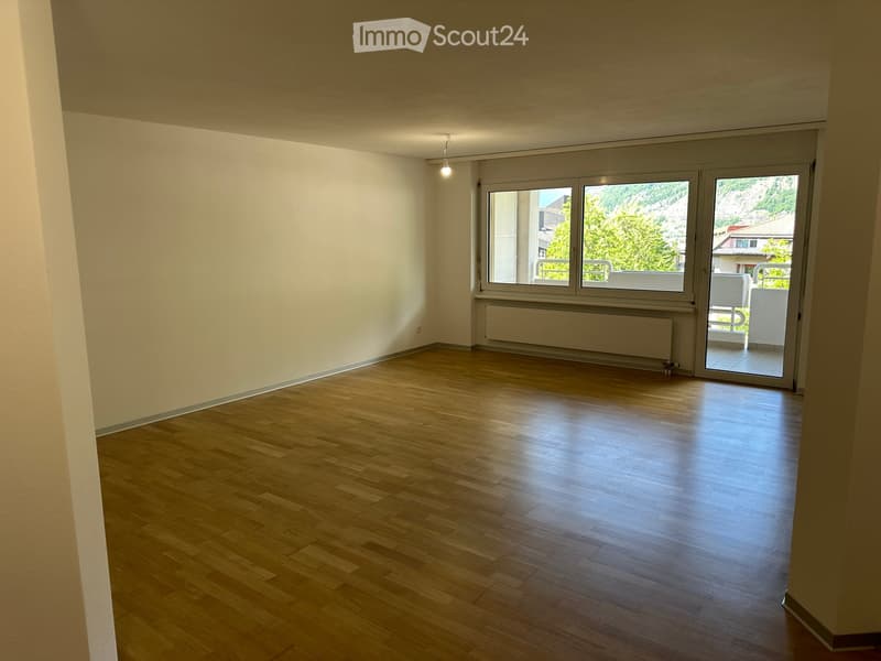 Empty room with wooden flooring, white walls, large windows, and a sliding glass door leading to a balcony.