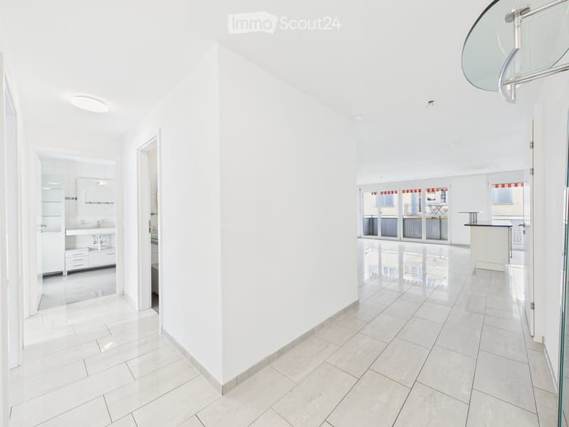 Bright, modern hallway with white walls and tiled flooring, leading to a bathroom and a room with large windows and a balcony view.