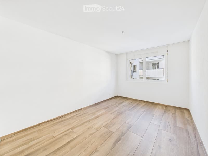 Empty room with white walls and wooden flooring, featuring a large window with a view of a building and balcony.
