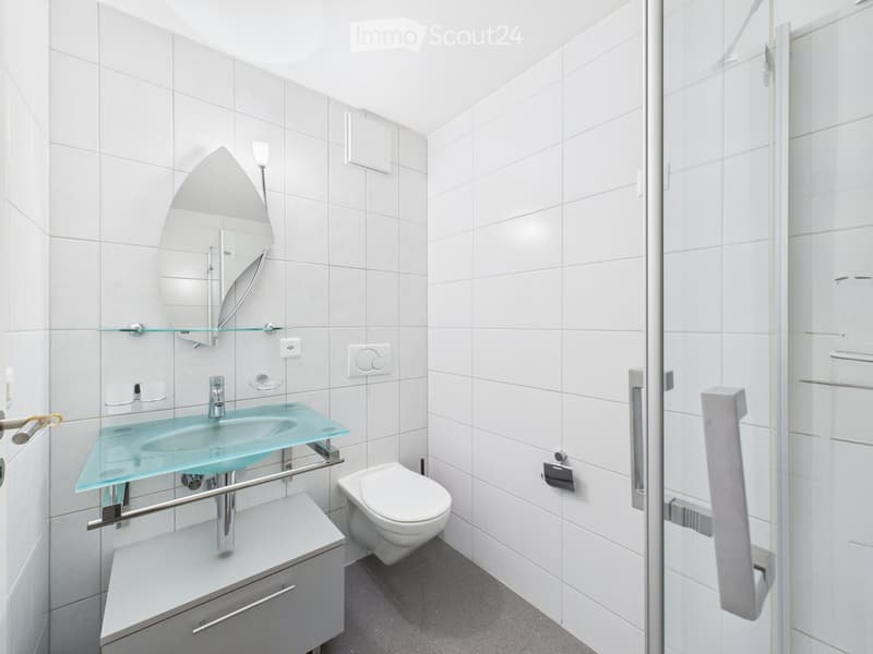 Modern bathroom with white tiled walls, a glass sink, a toilet, a large mirror, and a glass shelf above the sink.