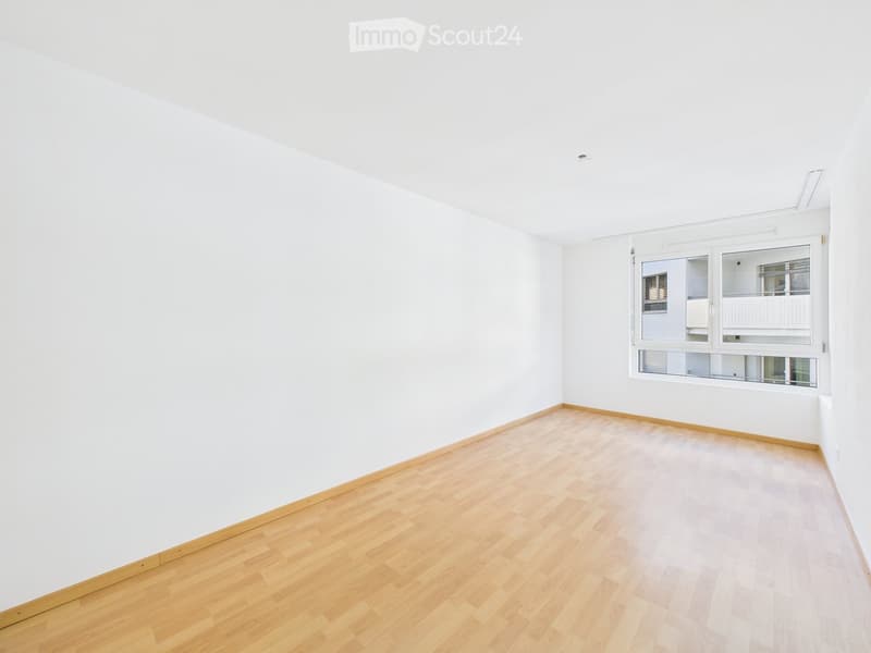 Empty room with white walls and light wooden flooring, featuring a large window with a view of a balcony and adjacent building.