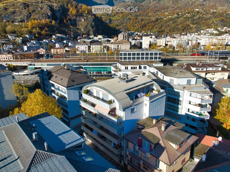 Aerial view of a multi-story residential building complex with modern architecture, surrounded by other buildings and a mountainous backdrop.