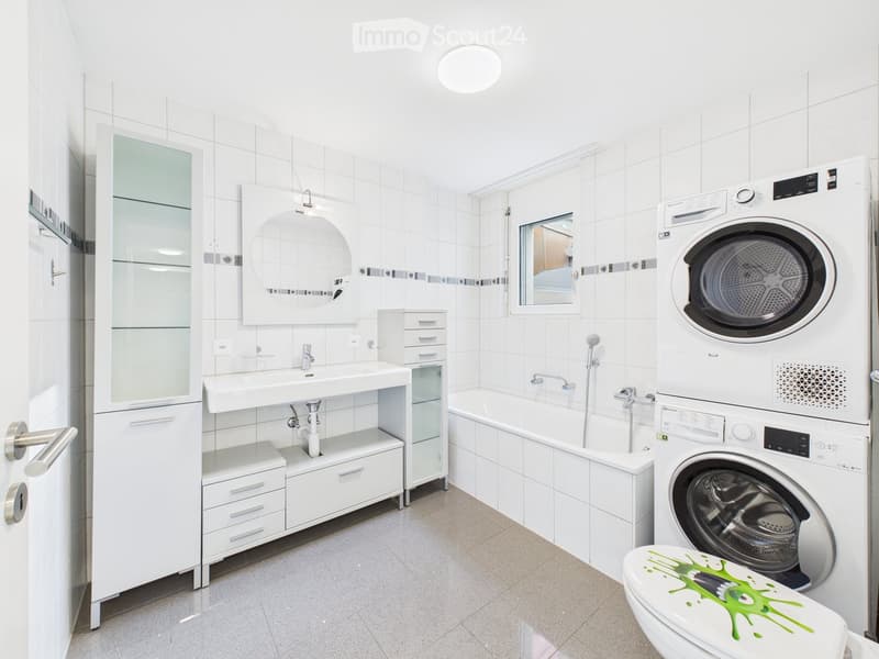 Modern bathroom with white tiles, a sink, bathtub, mirror, cabinet, washing machine, and a toilet with a green splatter design.