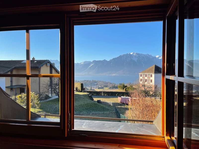 View from a window showing a scenic landscape with mountains, a lake, and surrounding houses.