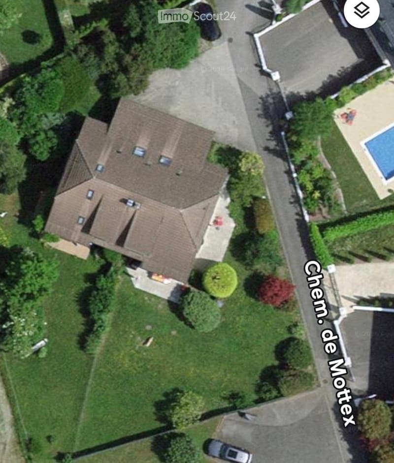 Aerial view of a two-story house with a tiled roof, surrounded by a well-maintained lawn and trees. A driveway leads to the house, and there is a swimming pool visible in the neighboring property.