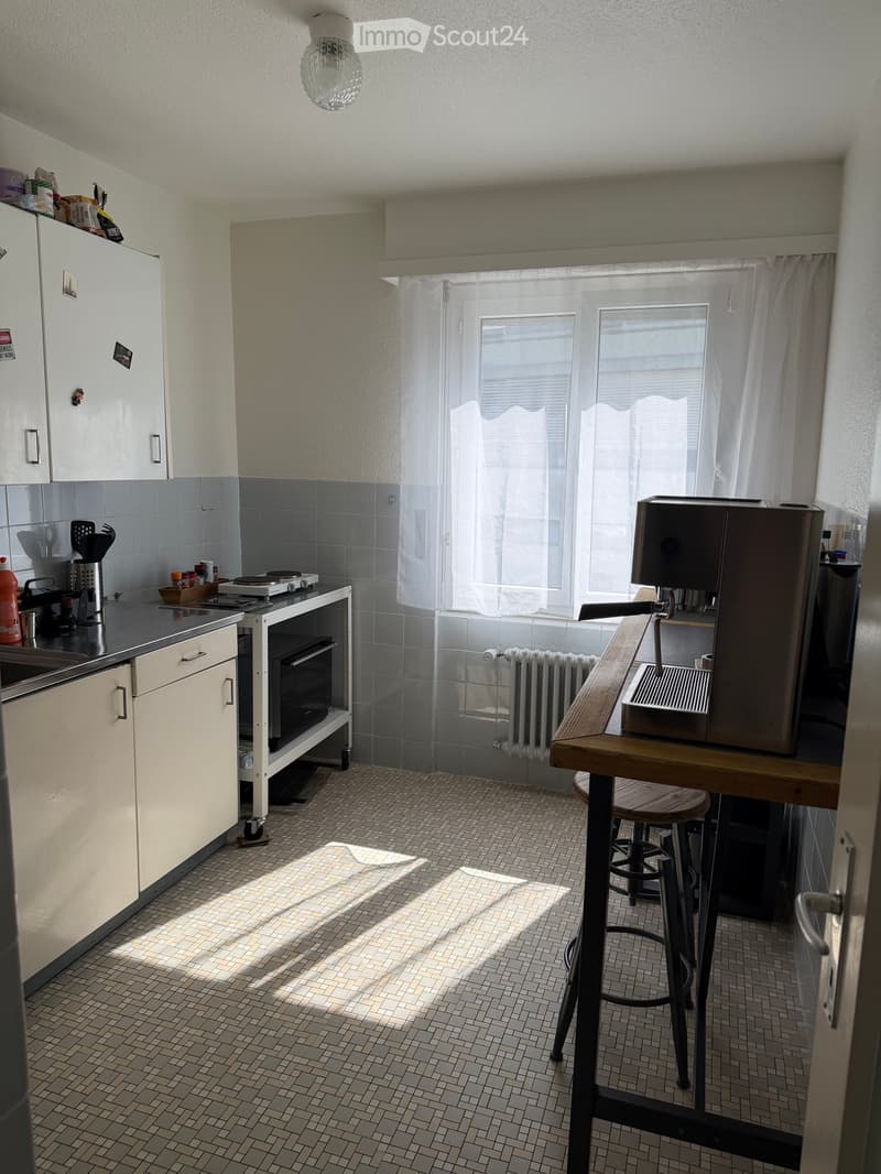 The kitchen features white cabinets, a tiled backsplash, and a tiled floor. A coffee machine sits on a wooden counter with a stool, and a small oven is positioned near the sink. Natural light streams in through a window with white curtains.