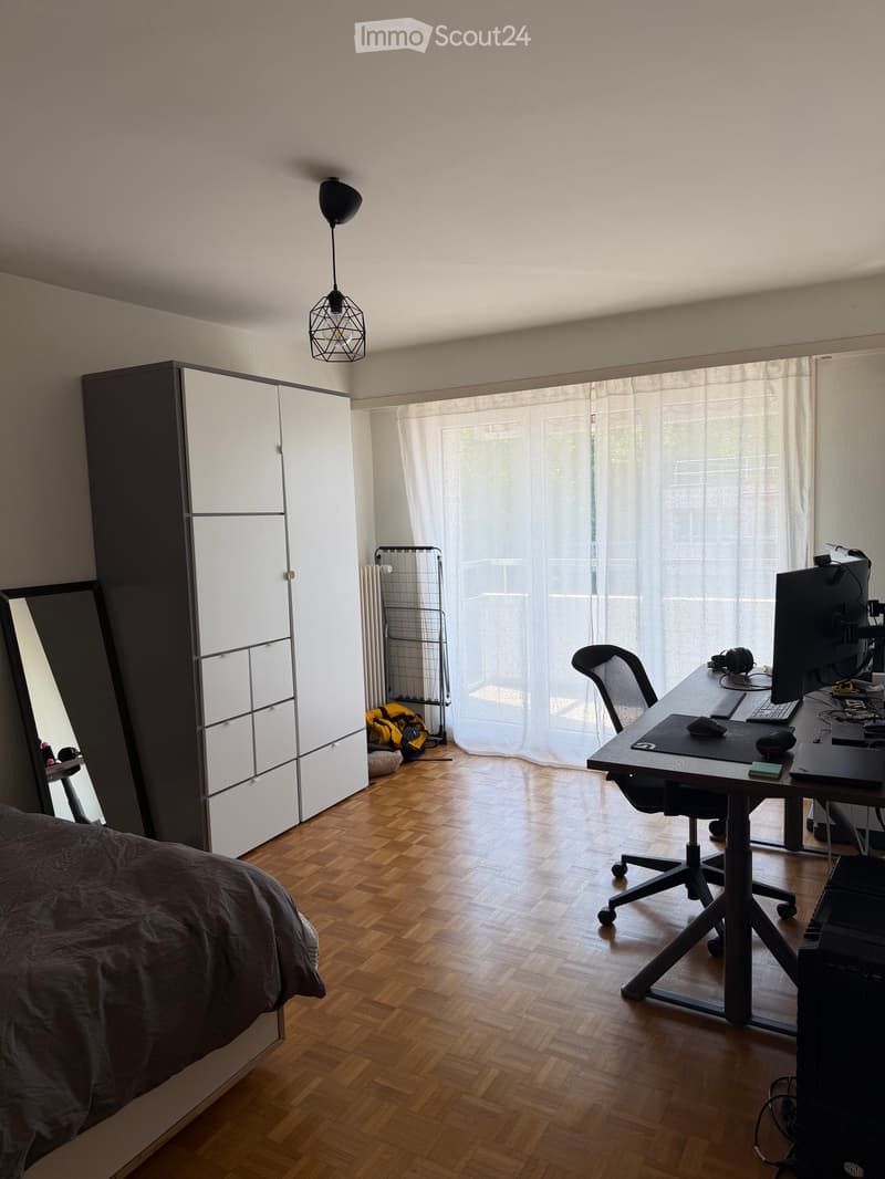 A well-lit room with a bed, desk, and wardrobe. The room features wooden flooring, a large window with white curtains, and a hanging geometric light fixture.